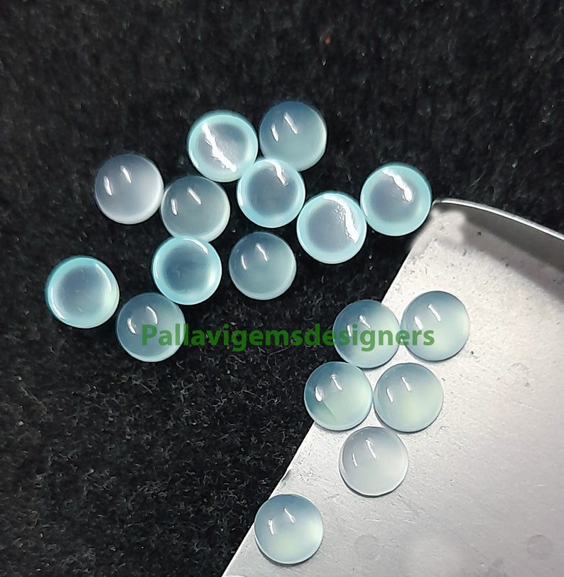 May include: A collection of 15 light blue round cabochon gemstones. The gemstones are smooth and polished, with a slightly translucent appearance. The gemstones are arranged in a loose pile on a black surface.
