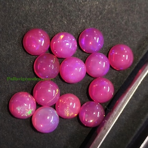 Puede incluir: Una colección de piedras preciosas de ópalo rosa redondas y pulidas. Los ópalos tienen una apariencia vibrante y translúcida con destellos de color internos. Las piedras están dispuestas sobre una superficie oscura, con una herramienta visible a la derecha.
