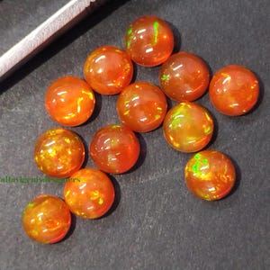 May include: A collection of round, orange-red opal gemstones with internal color flashes. The opals are scattered on a dark surface, with a pair of tweezers visible in the upper left corner. The gemstones have a polished, glossy appearance.