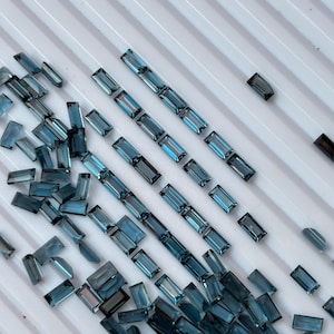May include: A collection of small, rectangular, baguette-cut blue gemstones arranged in neat rows and scattered clusters. The stones exhibit a deep, translucent teal-blue hue with faceted surfaces that reflect light, creating a uniform appearance.