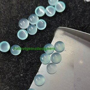 May include: A collection of 15 round, light blue cabochon gemstones. The gemstones are smooth and polished, with a translucent appearance. They are arranged on a silver surface and a black surface.