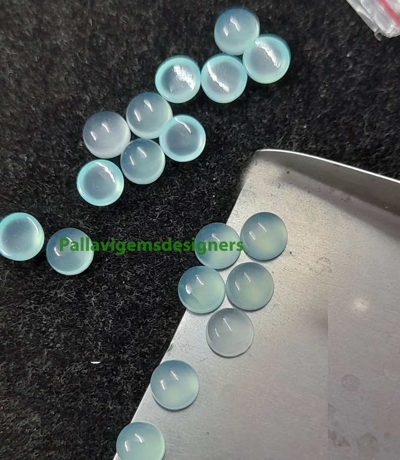 May include: A collection of 15 round, light blue cabochon gemstones. The gemstones are smooth and polished, with a translucent appearance. They are arranged on a silver surface and a black surface.