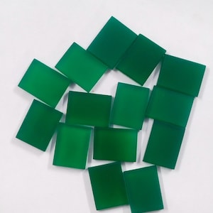 May include: A collection of square, emerald green glass tiles arranged on a white surface. The tiles are evenly cut and have a smooth, polished appearance. The arrangement is scattered, with some tiles overlapping.