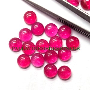 May include: A collection of round, polished, pink-red gemstones. The gems are arranged on a white surface, with a pair of tweezers visible in the background. The text "PALLAVIGEMSDESIGNERS" is printed in green across the image.