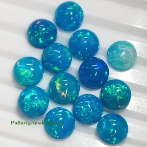Pode incluir: Uma coleção de pedras preciosas de opala redondas e polidas em tons de azul e verde-azulado. As opalas têm uma qualidade brilhante e iridescente, com flashes de verde e rosa. As pedras preciosas estão dispostas sobre uma superfície branca.