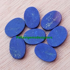 May include: Seven oval-shaped lapis lazuli cabochons, each with a deep blue colour and flecks of gold pyrite. The stones are arranged in a semi-circle on a wooden surface.