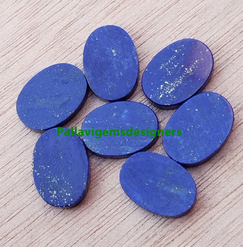 May include: Seven oval-shaped lapis lazuli cabochons, each with a deep blue colour and flecks of gold pyrite. The stones are arranged in a semi-circle on a wooden surface.