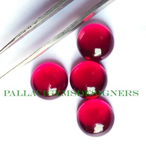 May include: Four round, red cabochon gemstones, each approximately 1 inch in diameter, are displayed on a white surface. The gemstones are smooth and shiny, with a slight variation in color from a deep red to a lighter red. A pair of silver tweezers is also visible in the image.