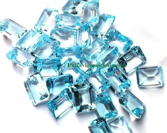 May include: A pile of light blue topazes cut into emerald shapes. The gemstones are faceted and have a clear, glassy appearance.