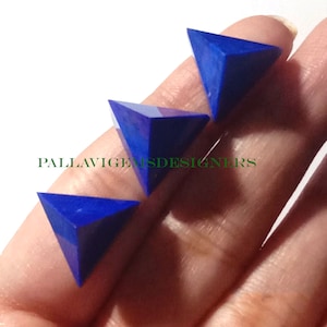 May include: Three blue lapis lazuli pyramid shaped gemstones. The gemstones are faceted and have a deep blue color with some white and purple inclusions. The gemstones are on a hand.