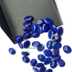 Puede incluir: Una colección de piedras preciosas de lapislázuli de forma ovalada, cada una de un color azul intenso y motas de pirita dorada. Las piedras están dispersas sobre una superficie blanca.