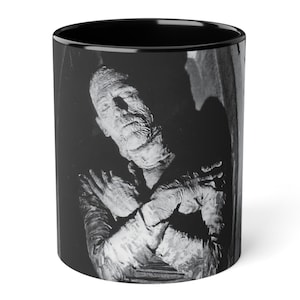 Puede incluir: Taza de cerámica en blanco y negro con una imagen clásica de una momia. La momia está representada con los brazos cruzados, sobre un fondo oscuro. La taza tiene un interior negro y un exterior blanco.