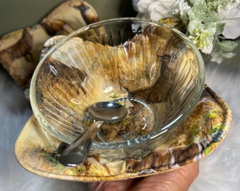 May include: A clear glass bowl with a decorative, earthy pattern, resting on a matching saucer. A small silver spoon sits inside the bowl. The bowl and saucer are held in a hand, with a blurred floral arrangement in the background.