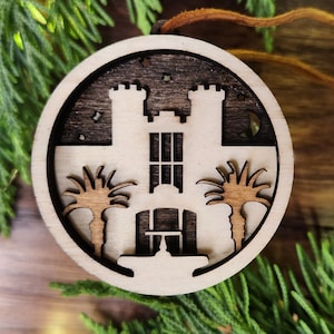 May include: A wooden ornament shaped like a castle with two palm trees on either side. The ornament is painted in shades of brown and black, and has a rustic look.