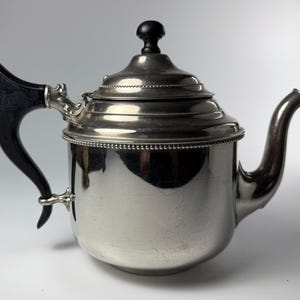 May include: A silver-colored teapot with a black handle and lid knob. The teapot has a curved spout and a decorative beaded trim around the top and bottom. The handle is curved and attached to the body with ornate metalwork.