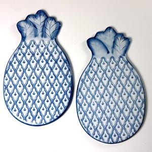 May include: Two ceramic dishes shaped like pineapples, featuring a white base with a blue lattice pattern and blue leaf details at the top. The dishes have a smooth, glossy finish and are arranged side by side on a plain white surface.