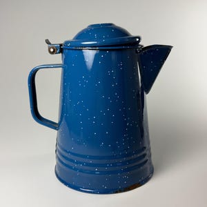 May include: A deep blue enamel coffee percolator featuring a white speckled pattern throughout the body. The vessel includes a matching lid with a metal hinge, a side handle, and a wide spout. The base shows signs of wear with visible rust.