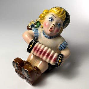 May include: A vintage ceramic figurine of a cherubic child playing an accordion. The figurine has blonde hair, blue eyes, and rosy cheeks. The child is wearing a hat and a striped shirt. A basket of flowers is behind the child.