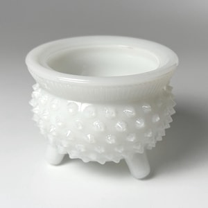 May include: A small, white milk glass bowl featuring a hobnail texture on the exterior. The bowl has a rounded body, a smooth flared rim, and stands on three short, sturdy legs. The surface is opaque and glossy with a distinct raised pattern.
