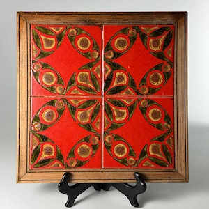 May include: A square wooden frame containing four ceramic tiles arranged in a grid. The tiles feature a vibrant red background with a repeating pattern of dark green and brown circular and leaf-shaped motifs across the surface.