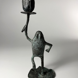 May include: A dark bronze-toned metal candlestick holder shaped like a standing frog on a lily pad base. The frog figure holds a long, slender stem topped with a flower-shaped cup designed to hold a candle, featuring a weathered, textured finish.