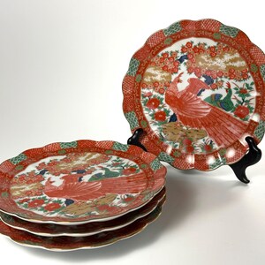 May include: A set of four round ceramic plates with scalloped edges, featuring a central red phoenix bird motif surrounded by green foliage and floral patterns. The border displays a red and green checkered design with gold accents.