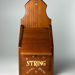 May include: A wall-mounted wooden string dispenser with a medium brown finish. The front panel features the word STRING printed in yellow, accompanied by small yellow floral and wheat-like decorative accents. It has a pointed top with a hole.