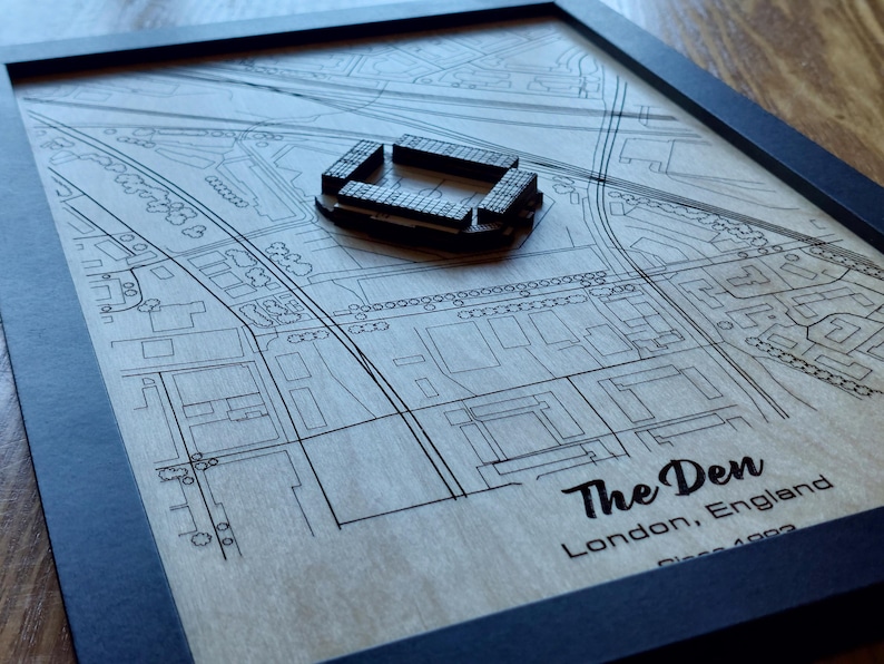 Pu&ograve; includere: Una mappa in legno di The Den, uno stadio a Londra, in Inghilterra. La mappa &egrave; incorniciata in nero e presenta un modello 3D dello stadio.