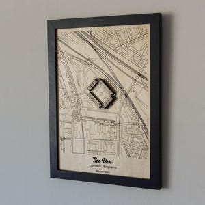 Pu&ograve; includere: Una mappa di legno incorniciata di The Den a Londra, in Inghilterra, con un ritaglio in legno 3D dello stadio. La mappa &egrave; beige con linee e testo neri. La cornice &egrave; nera.