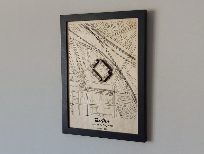 Pu&ograve; includere: Una mappa di legno incorniciata di The Den a Londra, in Inghilterra, con un ritaglio in legno 3D dello stadio. La mappa &egrave; beige con linee e testo neri. La cornice &egrave; nera.