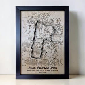 May include: A framed wall art piece featuring a topographic map of the Mount Panorama Circuit in Bathurst, New South Wales, Australia. The map includes a raised black outline of the race track over a beige background with printed text.