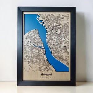 Puede incluir: Un mapa de madera en 3D enmarcado de Liverpool, Reino Unido, con una cuadrícula de calles cortada con láser en tonos de madera clara sobre una incrustación de agua azul vibrante. El marco negro rodea el mapa, que incluye el texto Liverpool y United Kingdom.