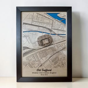 May include: A framed 3D wooden map of Old Trafford in Greater Manchester, England, established 1910. The light wood map features laser-cut street details, a raised stadium model, and blue water accents, all enclosed in a black rectangular frame.