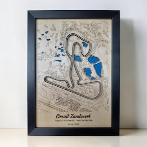 May include: A framed wooden map of the Circuit Zandvoort in North Holland, Netherlands. The map features a layered design with a light wood finish, blue cutouts representing water, and black track lines. Text reads: Circuit Zandvoort, North Holland, Netherlands, Since 1948.