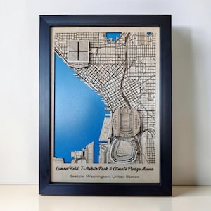 May include: A framed 3D wooden map of Seattle, Washington, featuring a blue water background, street grids, and raised models of Lumen Field, T-Mobile Park, and Climate Pledge Arena. The text reads Lumen Field, T-Mobile Park & Climate Pledge Arena.