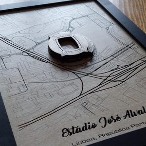 Op de afbeelding: Ingelijst kunstwerk met een gedetailleerde kaart en een 3D-stadionmodel. De kaart is in zwart-wit, met het stadion in het midden. De lijst is zwart en de tekst "Estádio José Alvalade" is zichtbaar.