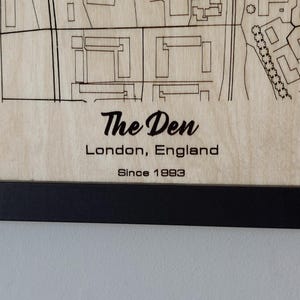Pu&ograve; includere: Un cartello in legno con il testo "The Den, Londra, Inghilterra, Since 1993" in lettere nere. Il cartello &egrave; incorniciato in nero.