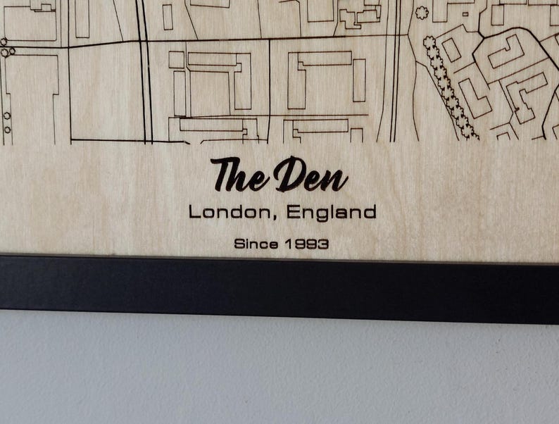 Pu&ograve; includere: Un cartello in legno con il testo "The Den, Londra, Inghilterra, Since 1993" in lettere nere. Il cartello &egrave; incorniciato in nero.