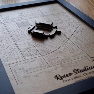Op de afbeelding: Een houten kaart met een 3D-model van het Reser Stadium in Corvallis, Oregon. De kaart is gelaserd met straatdetails en contouren van gebouwen. De lijst is zwart en het stadionmodel is verhoogd.