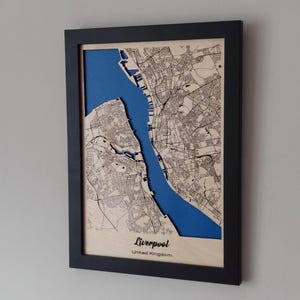 Pu&ograve; includere: Una mappa di legno incorniciata di Liverpool, Regno Unito, con un ritaglio blu del fiume Mersey. La mappa &egrave; tagliata al laser e presenta un design dettagliato della mappa stradale.