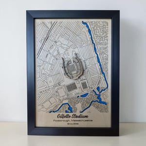 May include: A framed wooden map of Gillette Stadium in Foxborough, Massachusetts, featuring a layered 3D design with engraved street lines and a blue water element. The text Gillette Stadium, Foxborough, Massachusetts, Since 2002 is printed below.