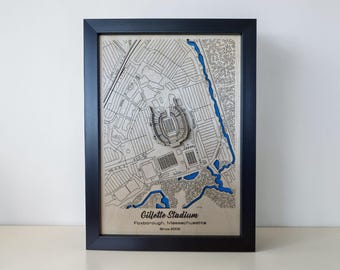 May include: A framed wooden map of Gillette Stadium in Foxborough, Massachusetts, featuring a layered 3D design with engraved street lines and a blue water element. The text Gillette Stadium, Foxborough, Massachusetts, Since 2002 is printed below.