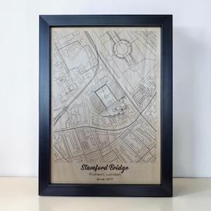 May include: A framed wooden wall art piece featuring a laser-engraved map of the Stamford Bridge stadium area in London. The light wood panel displays black street lines and stadium details, with the text Stamford Bridge, Fulham, London, Since 1877.