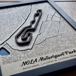 Puede incluir: Obra de arte en madera enmarcada del NOLA Motorsports Park en Avondale, Luisiana. El diseño de la pista está cortado con láser y en capas, creando un efecto 3D. La obra de arte está enmarcada en un marco negro, mostrando el diseño detallado.