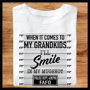 Peut inclure: T-shirt blanc avec un imprimé graphique noir et blanc qui dit "Quand il s'agit de mes petits-enfants, je vais sourire dans mon portrait judiciaire". Le graphique comprend également une photo de style portrait judiciaire avec le texte "Police Dept. 438798 FAFO".