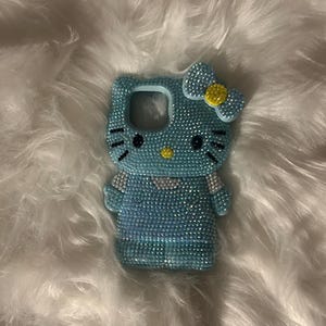 K&ouml;nnte beinhalten: Eine hellblaue Handyh&uuml;lle in Form einer Zeichentrickkatze, bedeckt mit glitzernden Strasssteinen. Die H&uuml;lle hat eine Schleife mit gelber Mitte und schwarzen Details f&uuml;r Augen und Schnurrhaare. Die H&uuml;lle liegt auf einer wei&szlig;en, flauschigen Oberfl&auml;che.