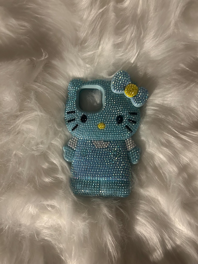 K&ouml;nnte beinhalten: Eine hellblaue Handyh&uuml;lle in Form einer Zeichentrickkatze, bedeckt mit glitzernden Strasssteinen. Die H&uuml;lle hat eine Schleife mit gelber Mitte und schwarzen Details f&uuml;r Augen und Schnurrhaare. Die H&uuml;lle liegt auf einer wei&szlig;en, flauschigen Oberfl&auml;che.