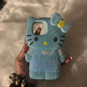 K&ouml;nnte beinhalten: Hellblaue Handyh&uuml;lle mit Hello Kitty-Design, vollst&auml;ndig mit glitzernden Strasssteinen besetzt. Die H&uuml;lle hat eine Schleife mit gelber Mitte und schwarzen Details. Die Handyh&uuml;lle wird in einer Hand gehalten.