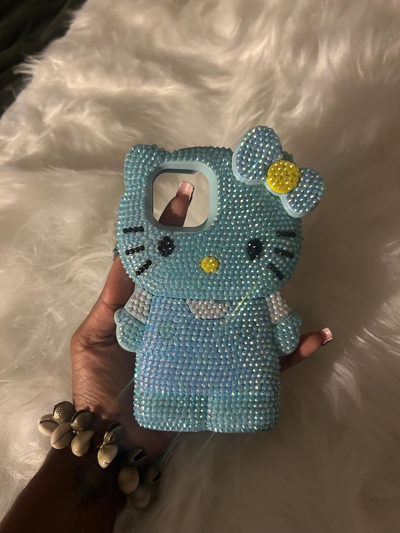 K&ouml;nnte beinhalten: Hellblaue Handyh&uuml;lle mit Hello Kitty-Design, vollst&auml;ndig mit glitzernden Strasssteinen besetzt. Die H&uuml;lle hat eine Schleife mit gelber Mitte und schwarzen Details. Die Handyh&uuml;lle wird in einer Hand gehalten.