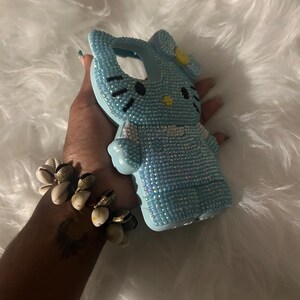 K&ouml;nnte beinhalten: Eine hellblaue Handyh&uuml;lle in Form einer Zeichentrickkatze, bedeckt mit kleinen, glitzernden Strasssteinen. Die Katze hat schwarze Augen und Schnurrhaare sowie eine gelbe Schleife. Die H&uuml;lle befindet sich vor einem flauschigen wei&szlig;en Hintergrund.